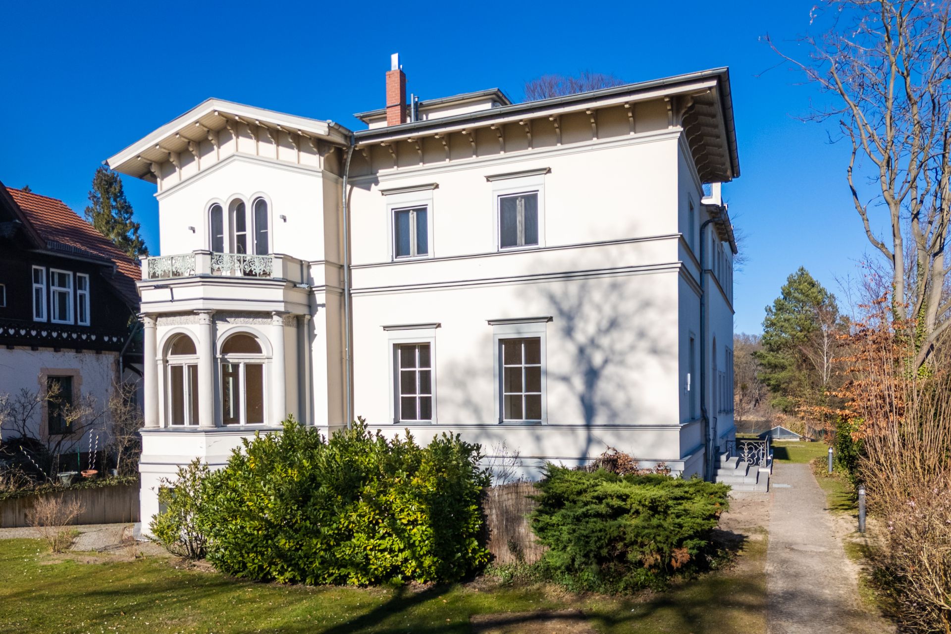 Helles, zweistöckiges Wohnhaus mit Balkon und großen Fenstern, umgeben von Garten und Bäumen.