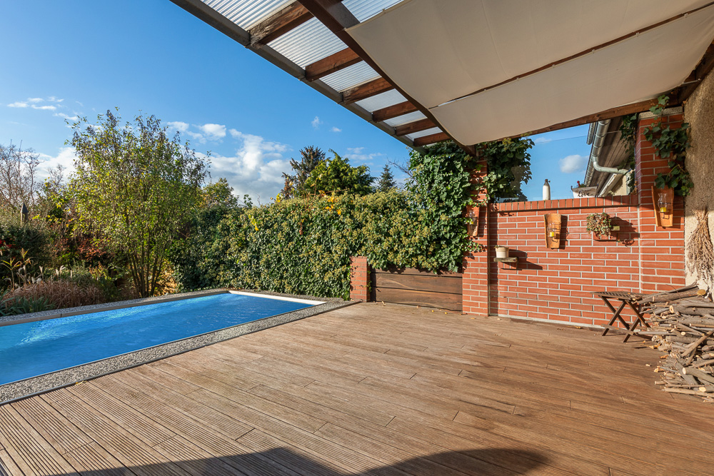 Überdachte Terrasse mit Holzfußboden, Pool und begrünter Garten im Hintergrund.