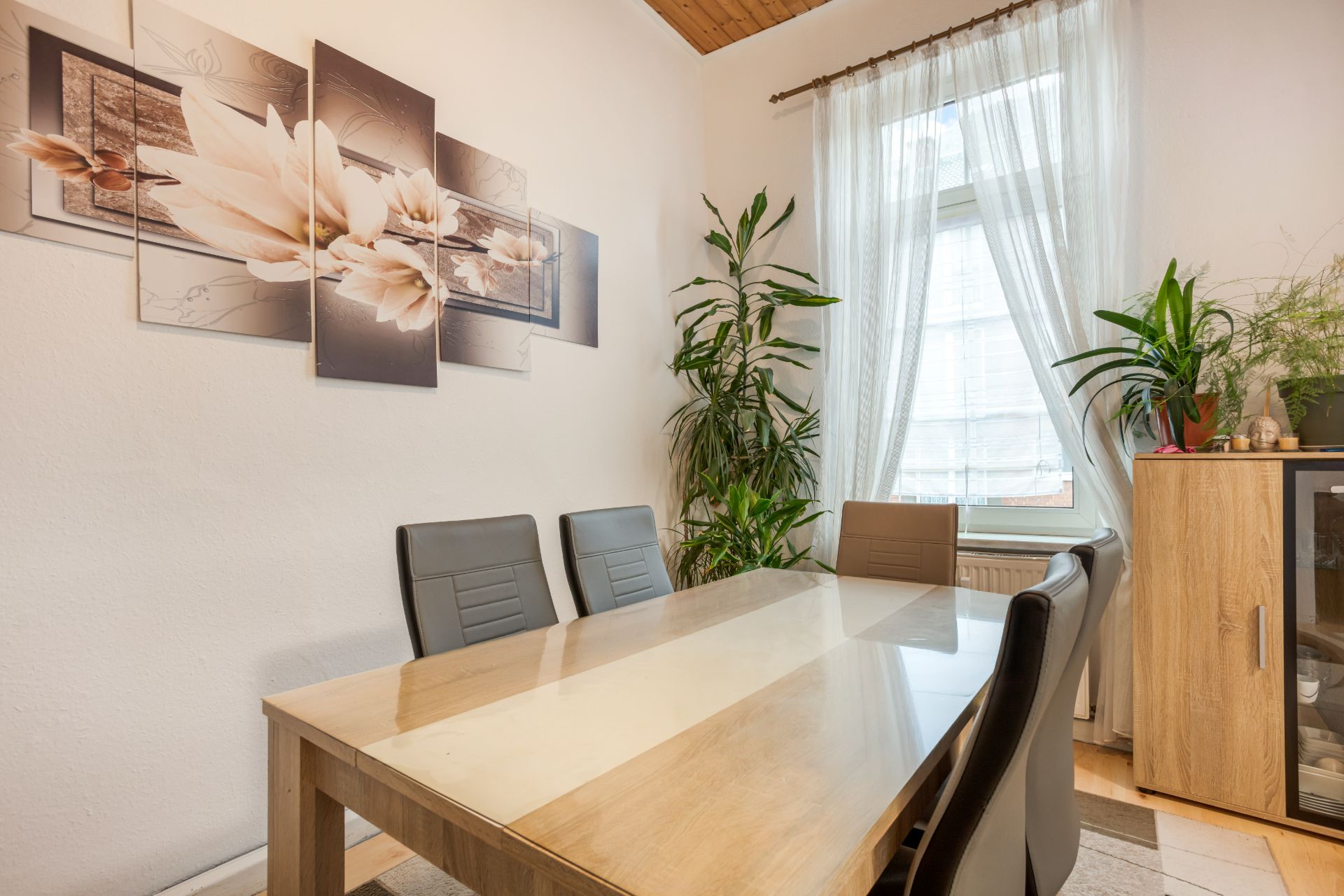 Esszimmer mit Holztisch, vier Stühlen, Pflanzen, Fenster mit Gardine und Wandbild mit Blumenmotiv.