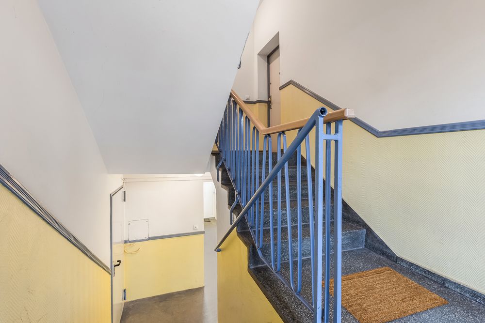 Treppenhaus mit Geländer, gelben Wänden und einer Fußmatte vor einer Wohnungstür.