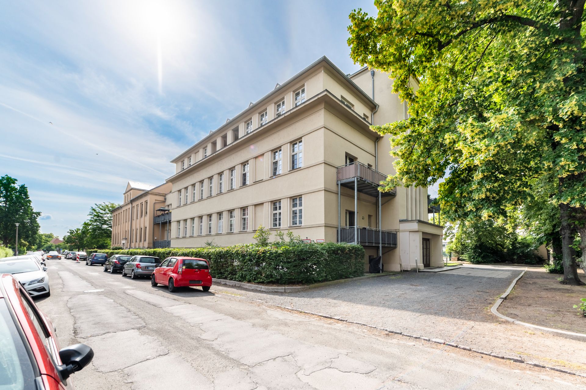 Mehrstöckiges Wohngebäude mit Balkonen an einer ruhigen Straße, Autos parken am Straßenrand.