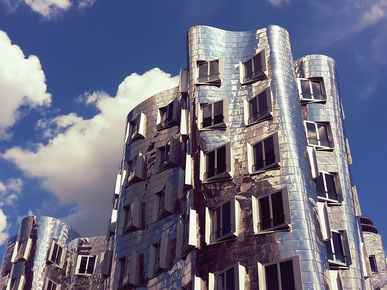 Modernes Wohngebäude mit wellenförmiger, silberner Fassade und vielen Fenstern vor blauem Himmel.