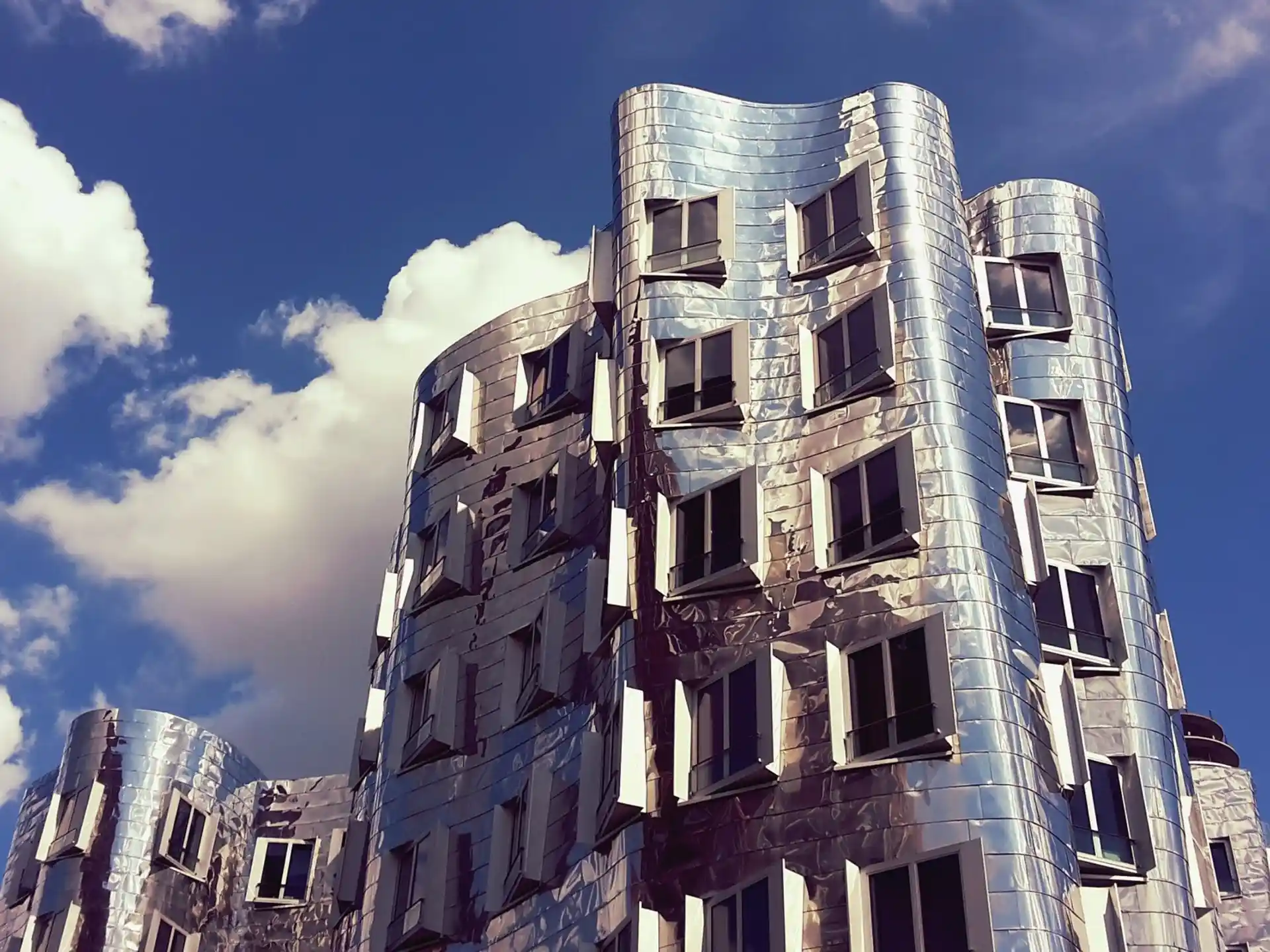 Modernes Wohngebäude mit wellenförmiger, silberner Fassade und vielen Fenstern vor blauem Himmel.