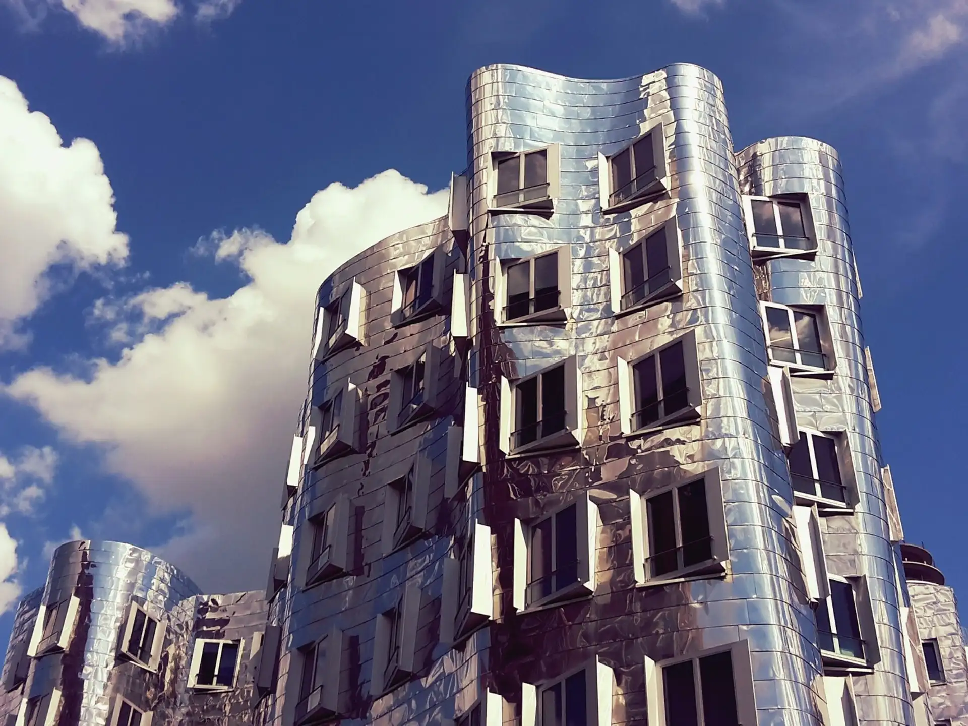 Modernes Wohngebäude mit wellenförmiger, silberner Fassade und vielen Fenstern vor blauem Himmel.