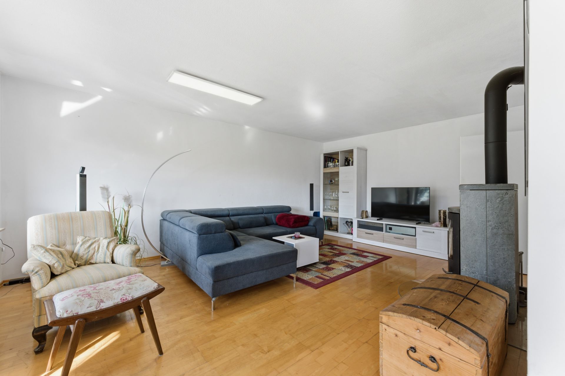 Helles Wohnzimmer mit blauem Sofa, Sessel, Holzofen, Fernseher und Holzboden.