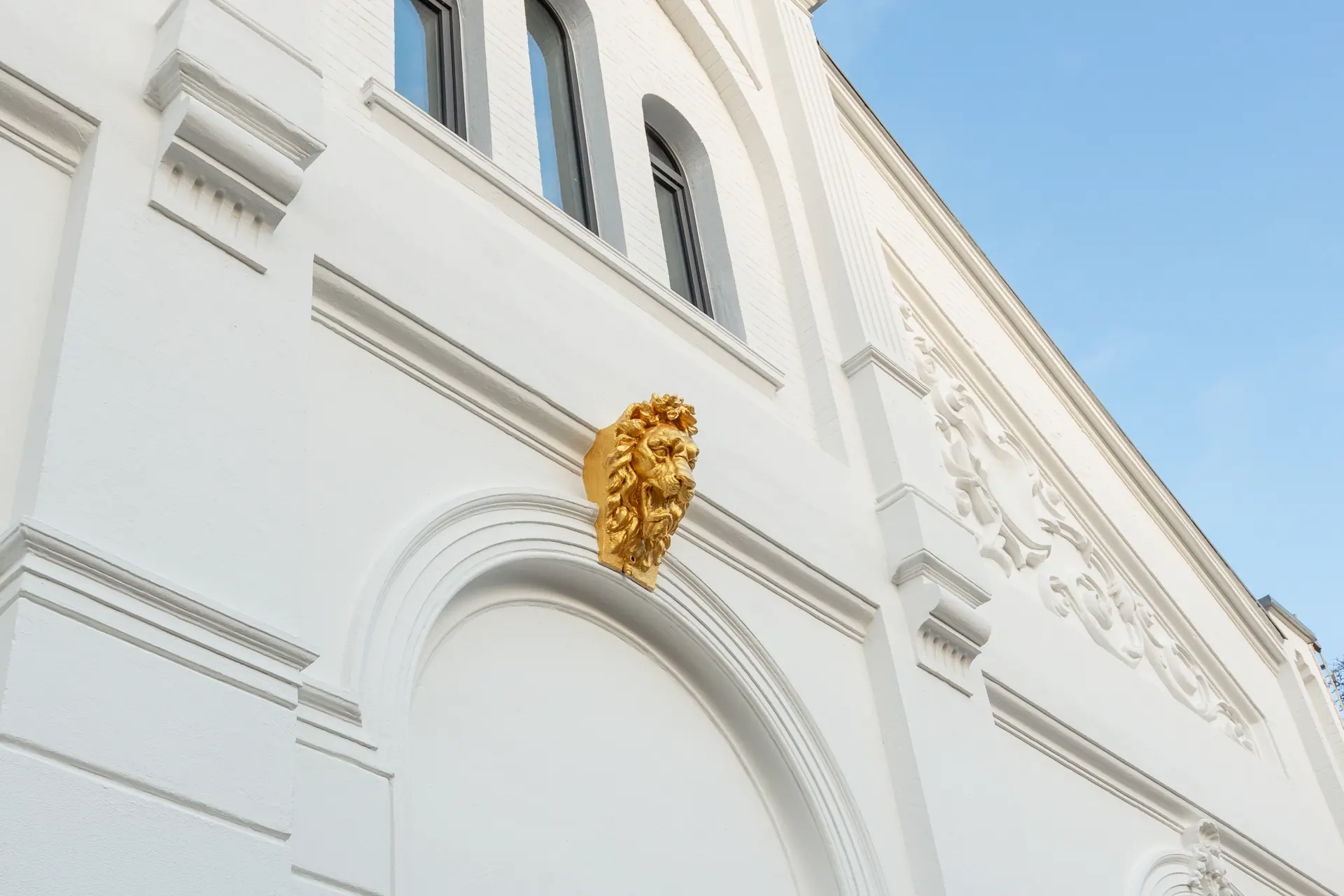 Weiße Fassade mit goldener Löwenkopf-Skulptur über einem Bogen und verzierten Details.