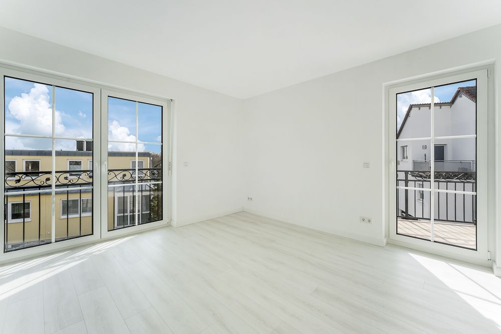Heller leerer Raum mit zwei großen Fenstern und einer Tür zu einem Balkon.