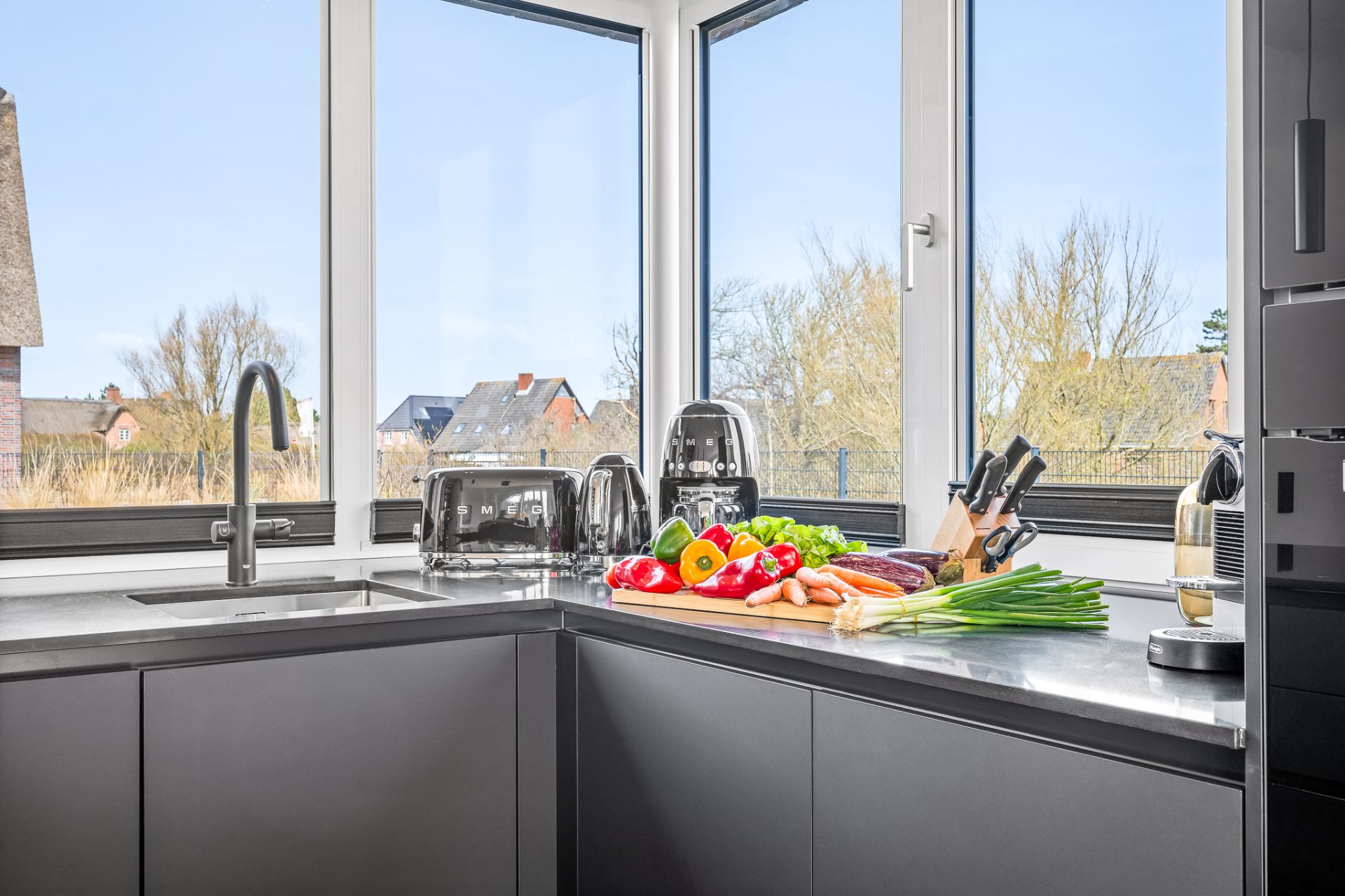 Moderne Küche mit großen Fenstern, Gemüse auf der Arbeitsplatte und Küchengeräten.