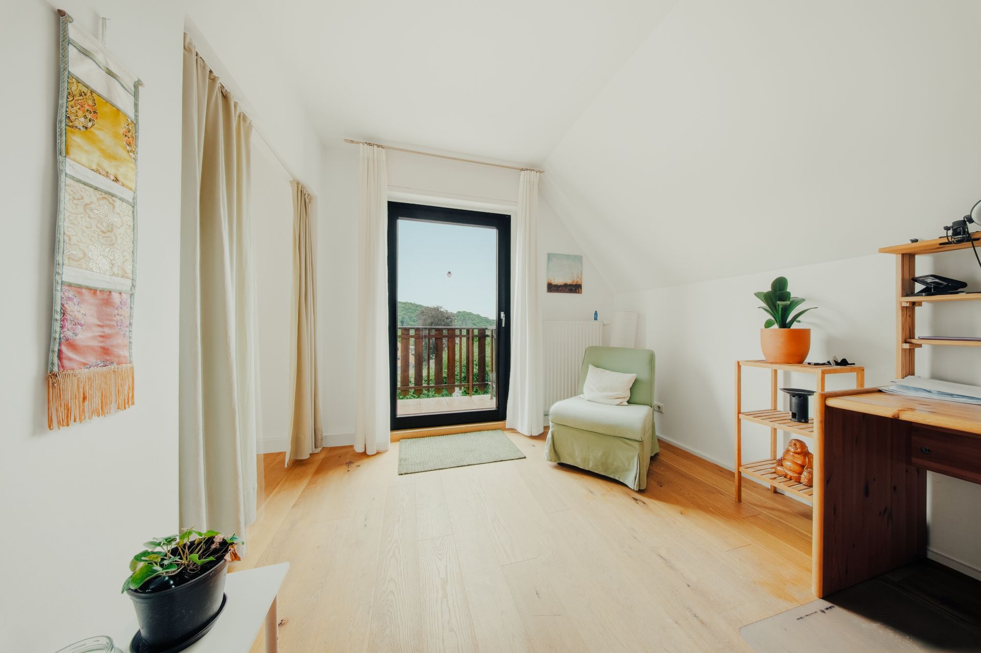 Helles Zimmer mit Holzfußboden, Sessel, Schreibtisch, Regal und Tür zum Balkon.