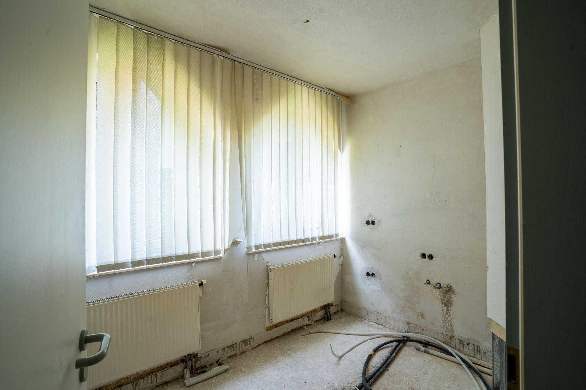 Leerer, renovierungsbedürftiger Raum mit zwei Fenstern, Heizkörpern und Lamellenjalousien.