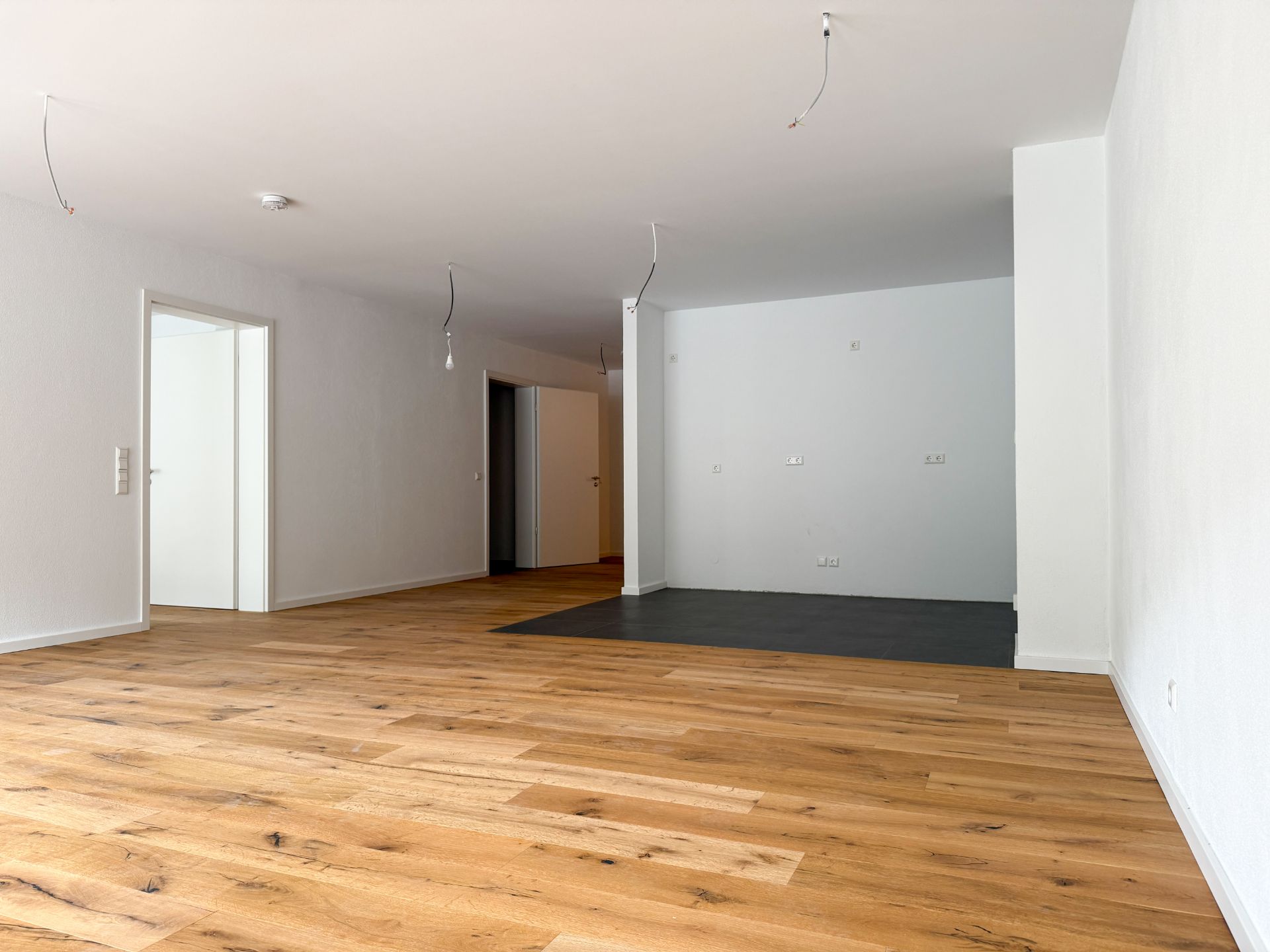 Leerer Raum mit Holzboden, weißen Wänden und offener Tür zu weiteren Zimmern.
