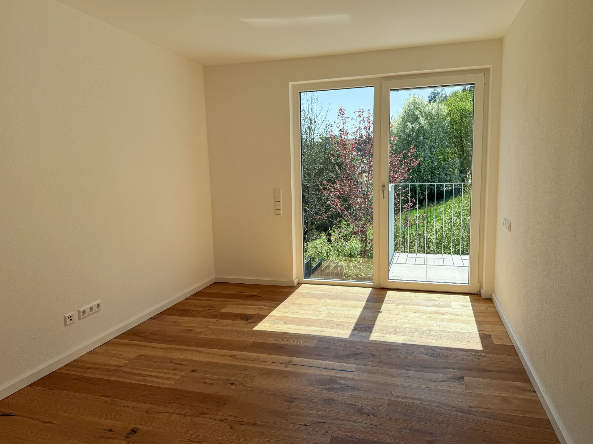 Heller leerer Raum mit Holzboden und großer Glastür zu einem kleinen Balkon mit Blick ins Grüne.