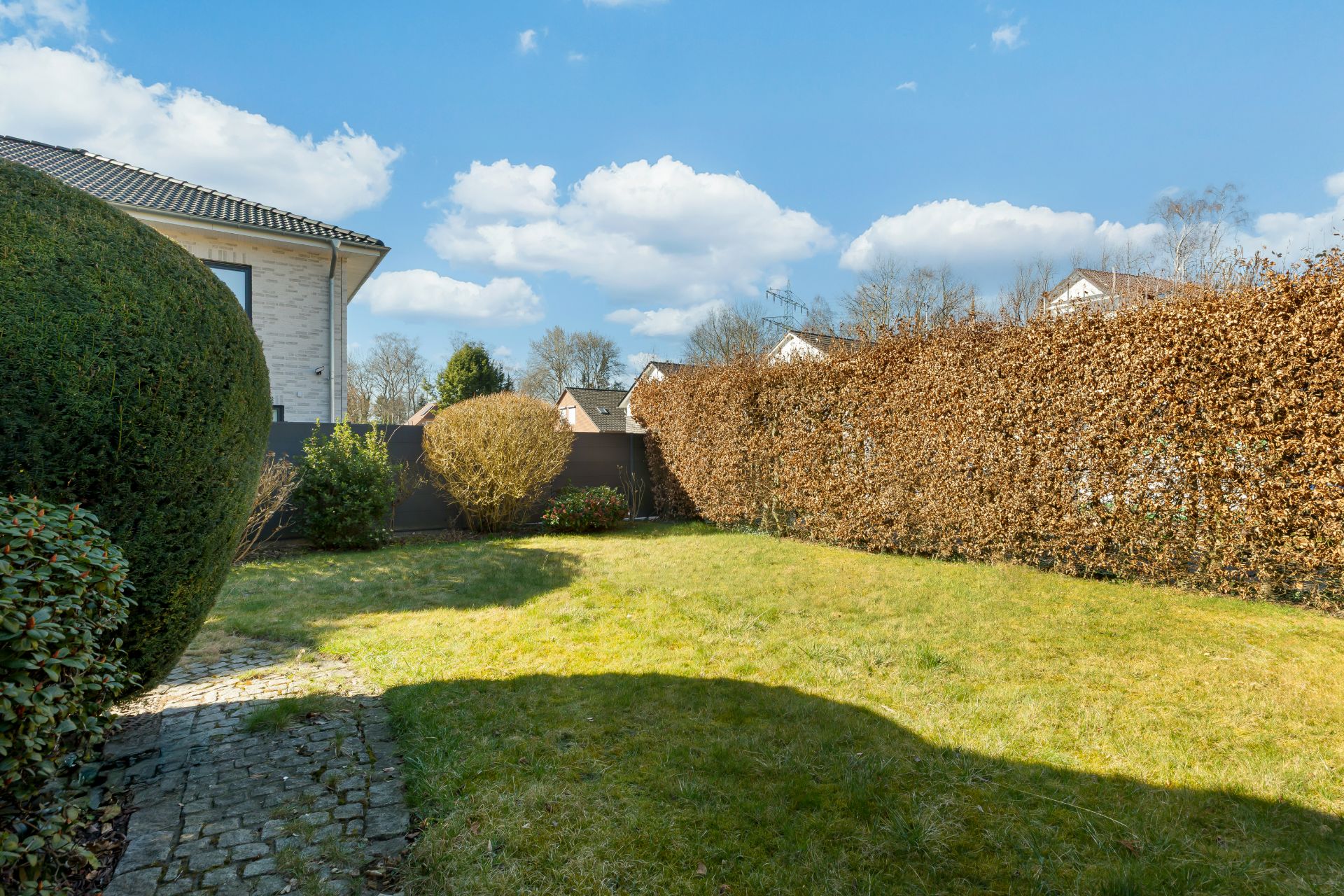 Grüner Garten mit Rasen, Hecke und gepflastertem Weg neben einem Wohnhaus bei sonnigem Wetter.