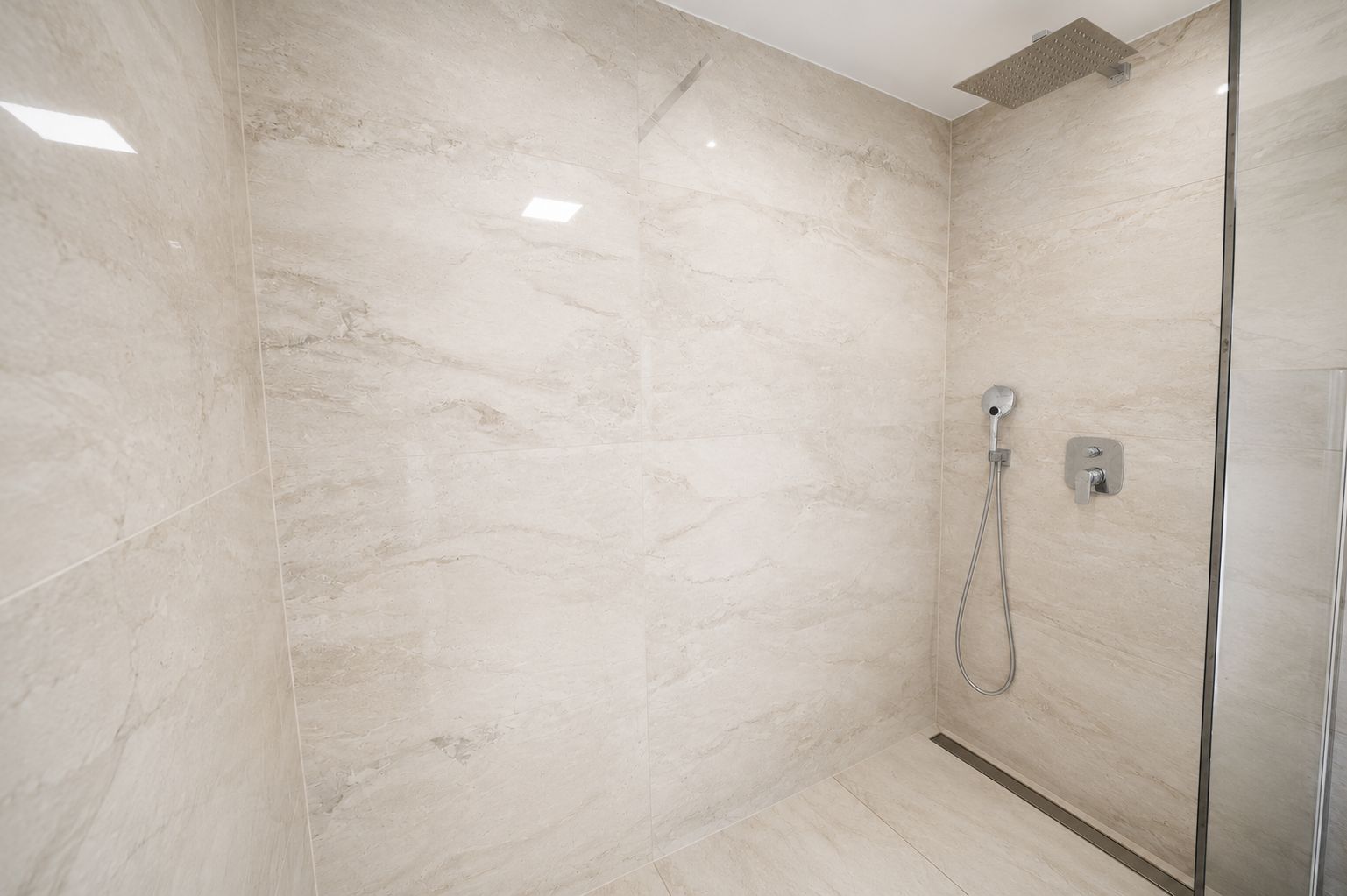 Moderne, begehbare Dusche mit großen, beigen Fliesen und Regenduschkopf.