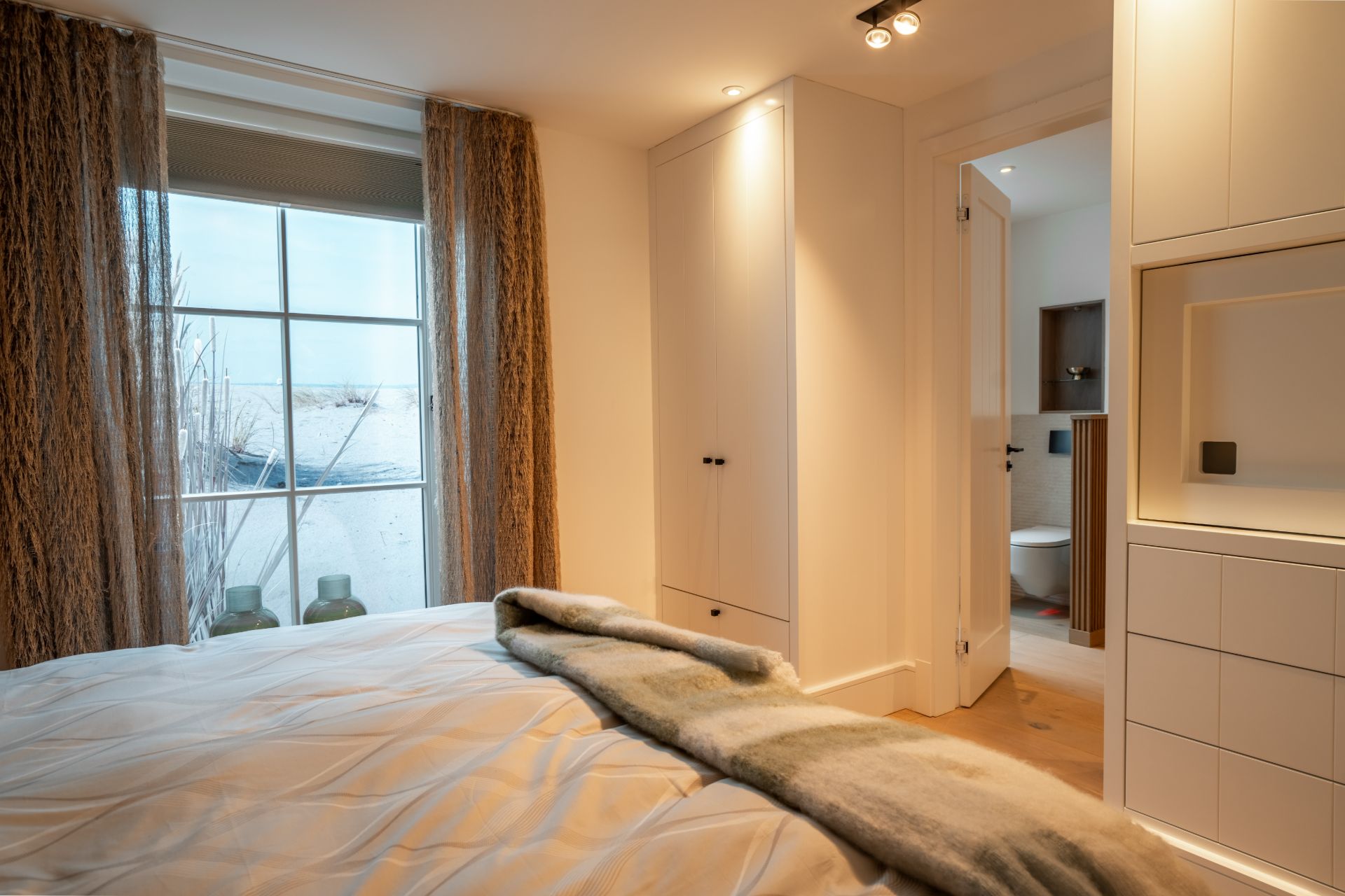 Schlafzimmer mit Doppelbett, großem Fenster, Gardinen und Blick ins Badezimmer.