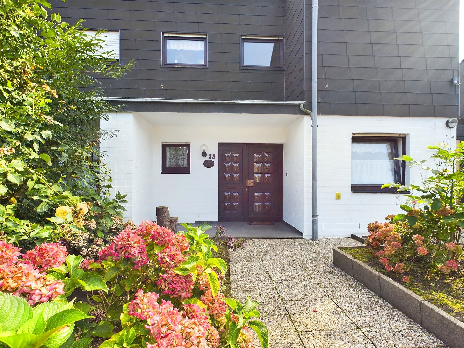 Eingang eines Wohnhauses mit brauner Tür, Blumenbeeten und gepflastertem Weg.