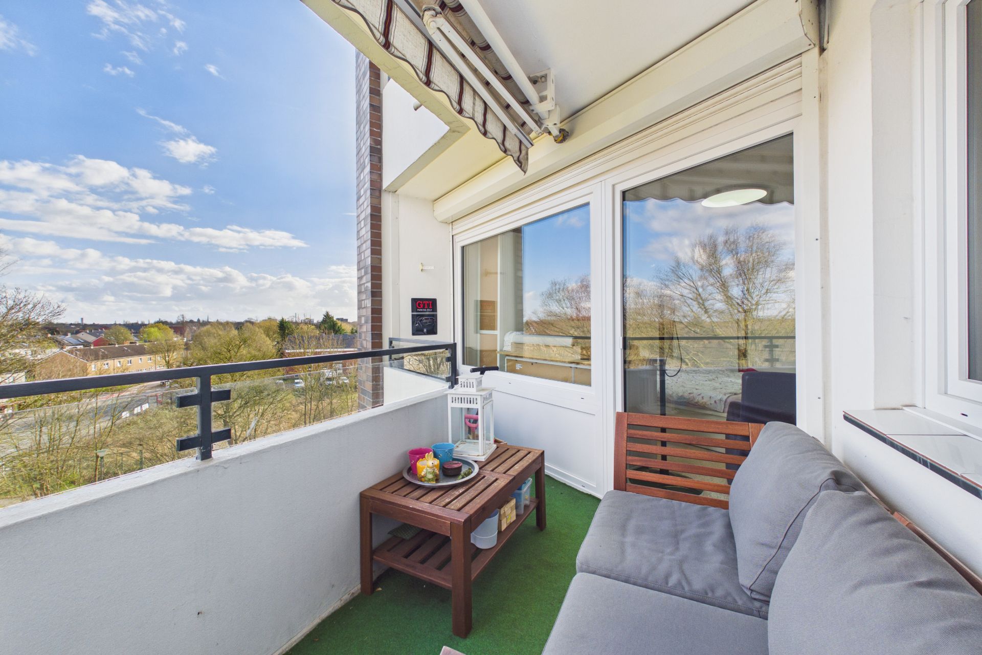 Balkon mit grauem Sofa, Holztisch und Ausblick auf Bäume und Häuser.