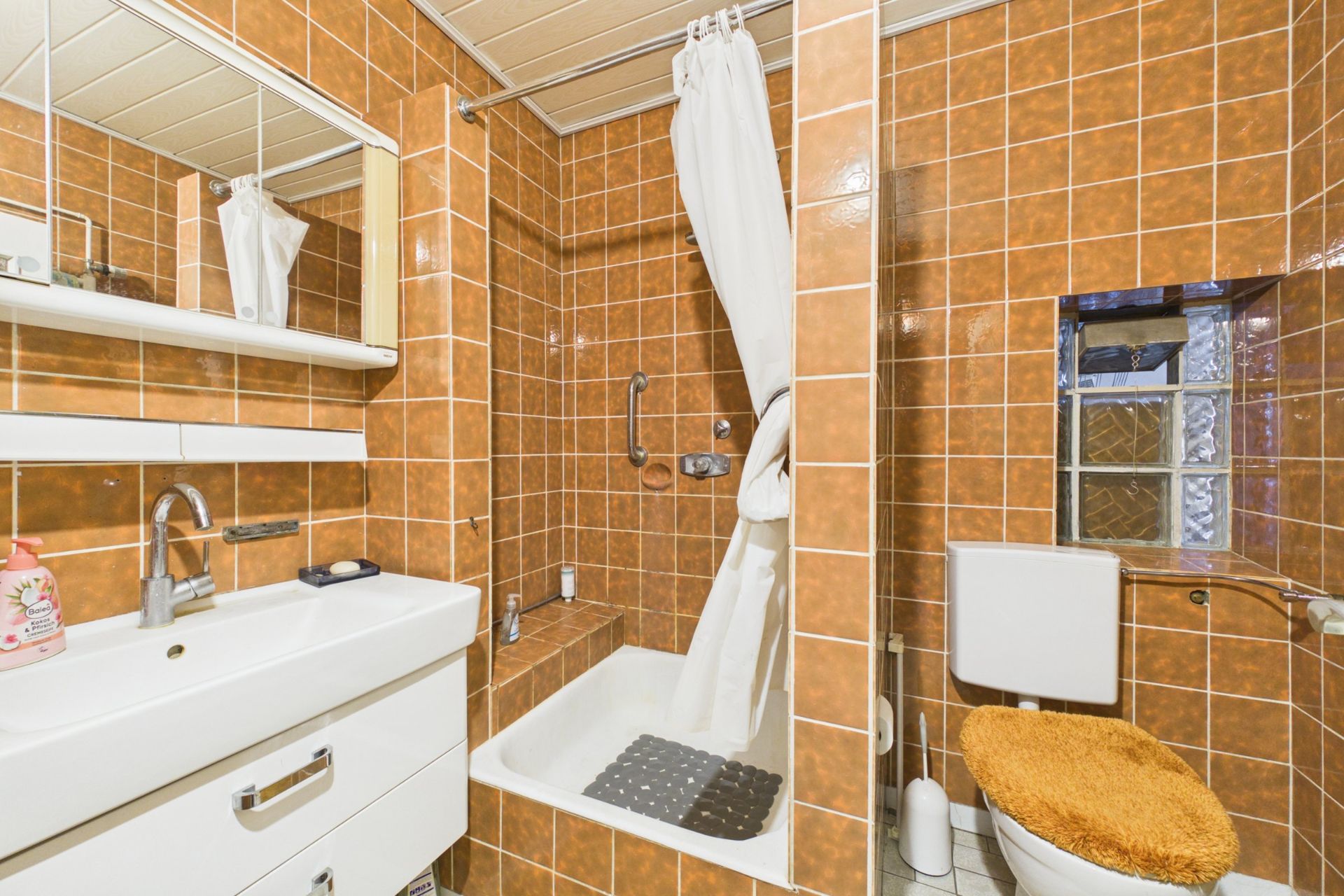 Badezimmer mit braunen Fliesen, Badewanne, Waschbecken und Toilette mit orangem Bezug.