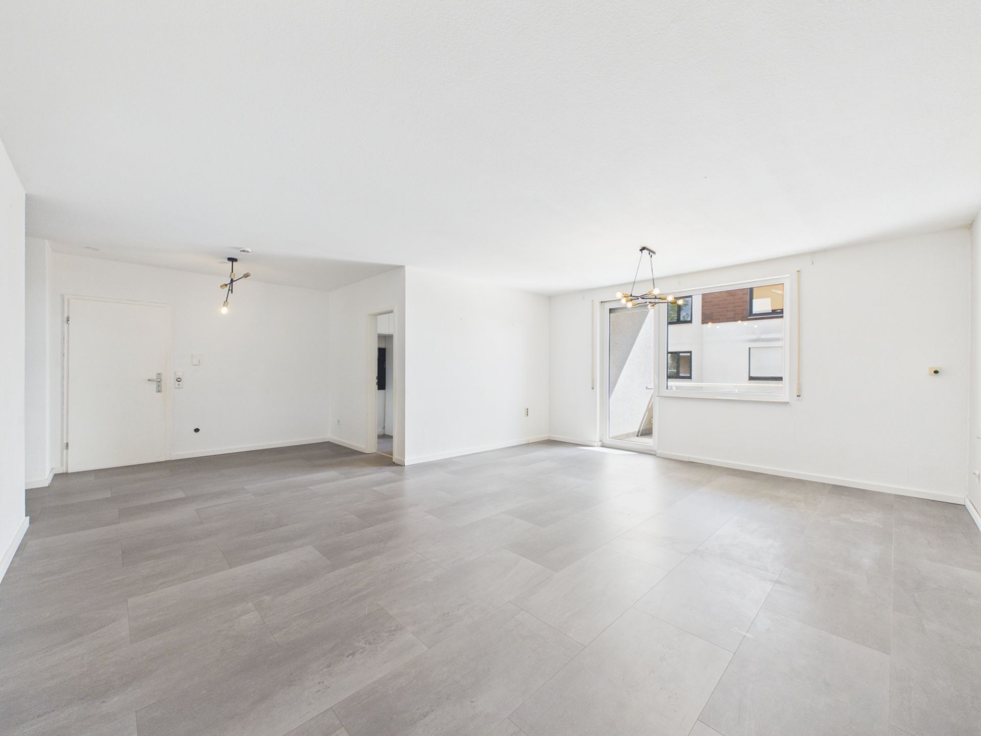 Helles, leeres Wohnzimmer mit grauem Boden, weißen Wänden und großen Fenstern.