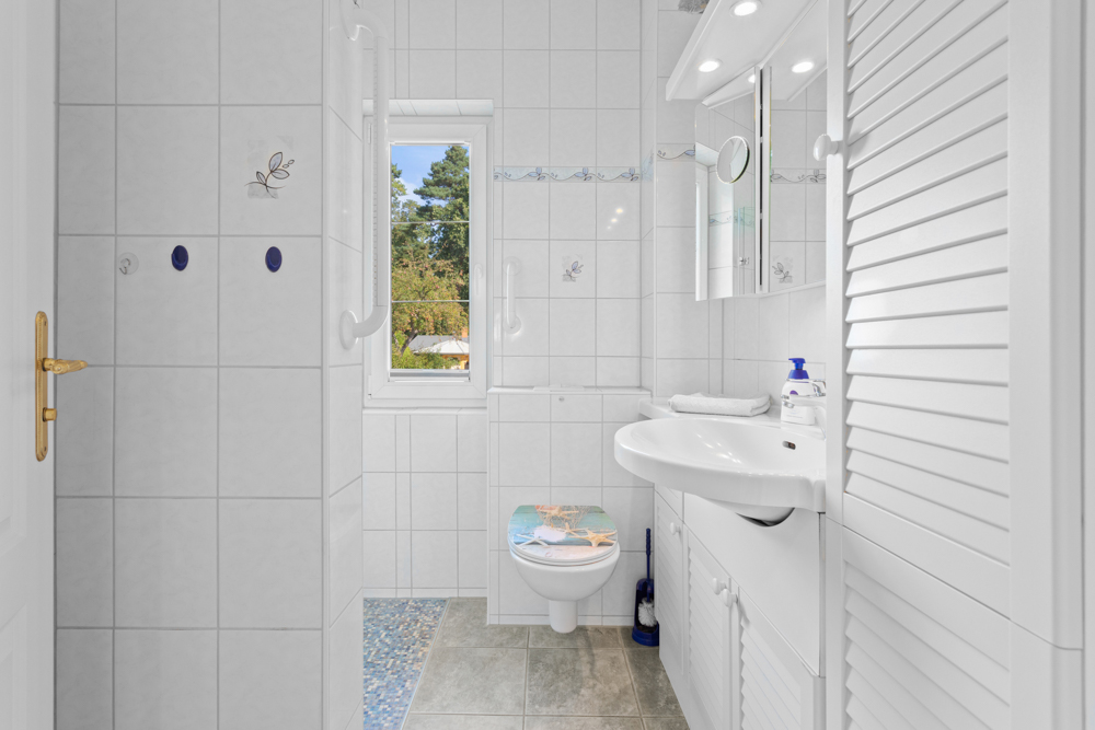 Helles Badezimmer mit WC, Waschbecken, Spiegel und Fenster, weiße Fliesen und Schränke.