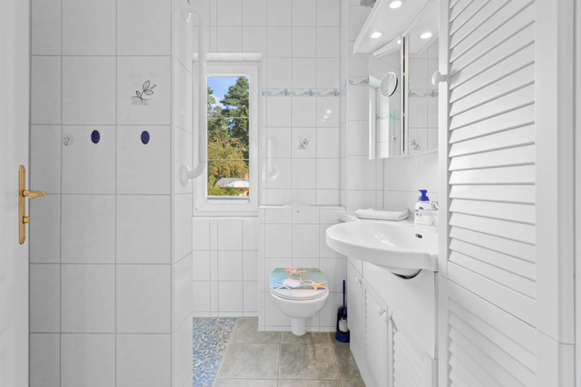 Helles Badezimmer mit WC, Waschbecken, Spiegel und Fenster, weiße Fliesen und Schränke.