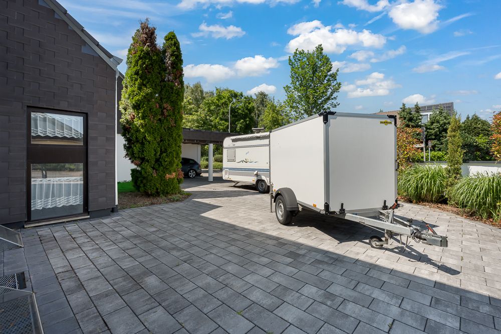 Wohnhof mit Pflastersteinen, Anhänger und Wohnwagen, daneben Bäume und modernes Gebäude.