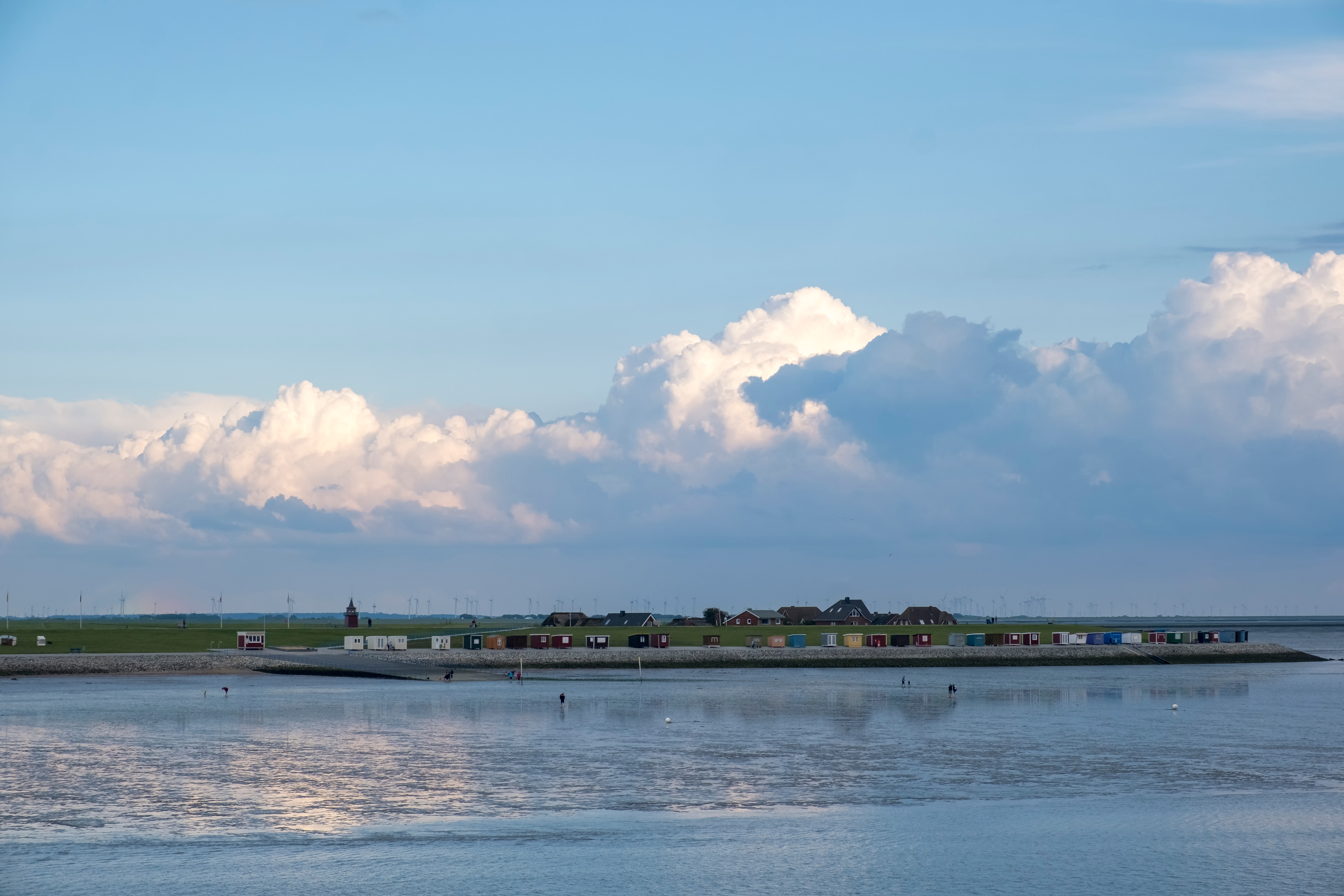 Kleine bunte Häuser stehen auf einer Wiese am Wasser unter einem bewölkten Himmel.