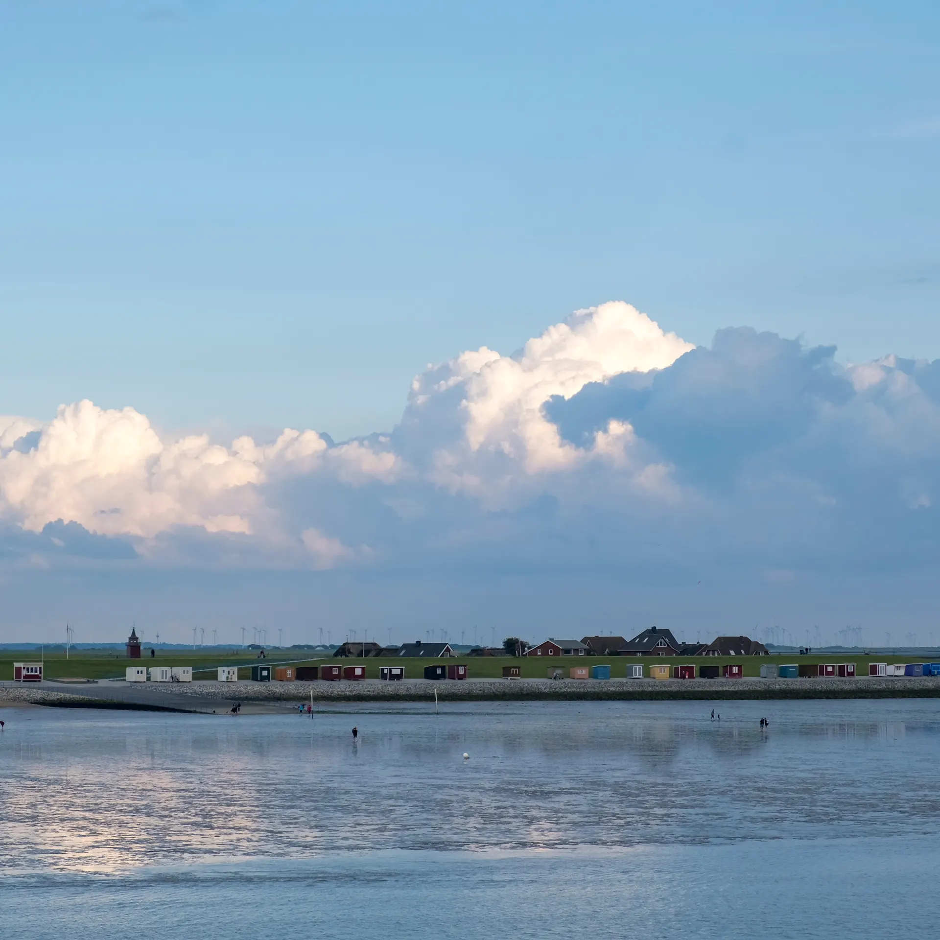 Kleine bunte Häuser stehen auf einer Wiese am Wasser unter einem bewölkten Himmel.