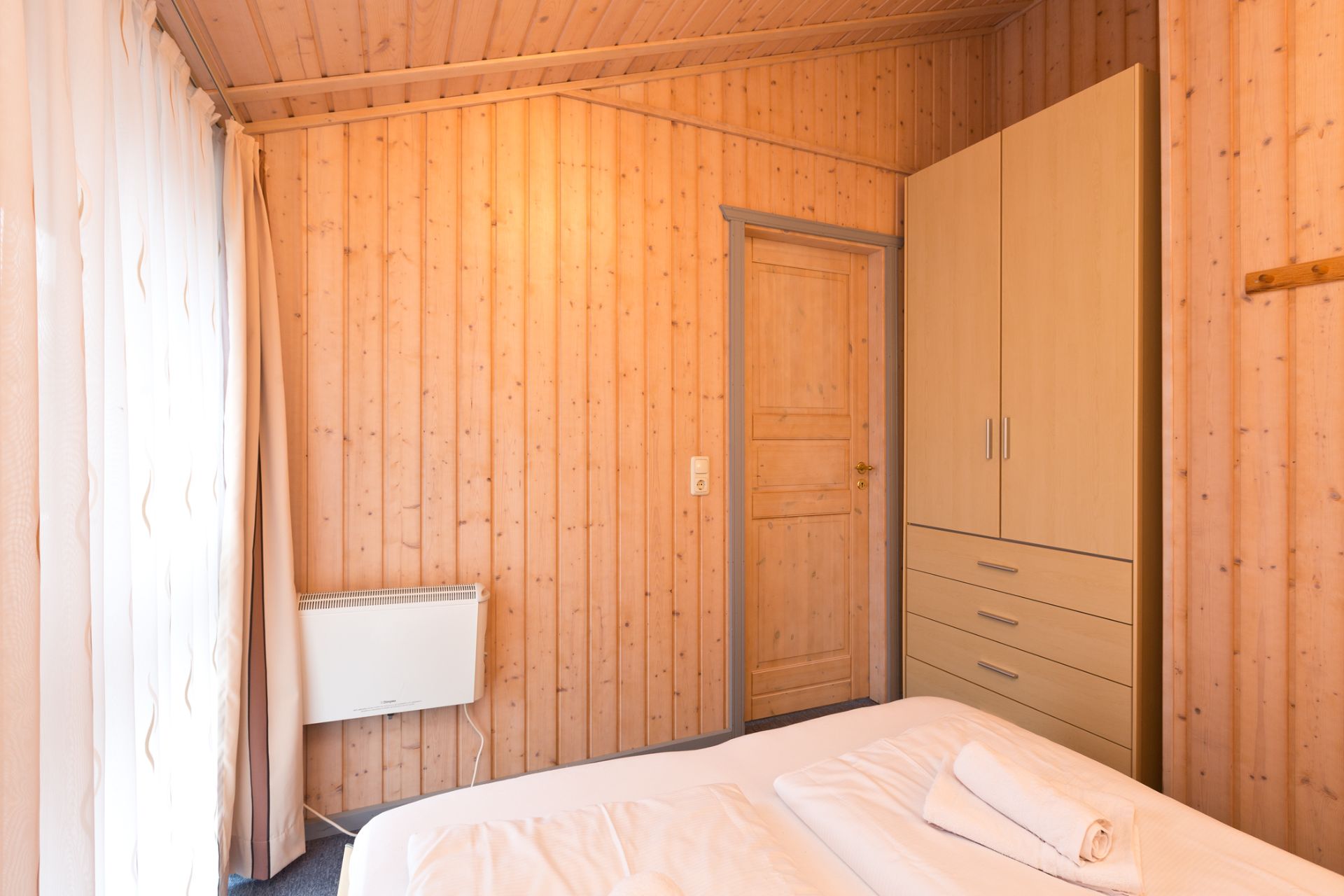 Kleines Schlafzimmer mit Holzwänden, einem Kleiderschrank, Bett und großem Fenster mit Vorhängen.