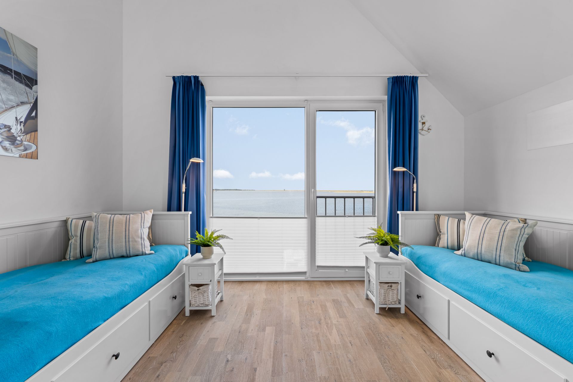 Zimmer mit zwei Einzelbetten, blauen Decken, Holzboden und großem Fenster mit Meerblick.