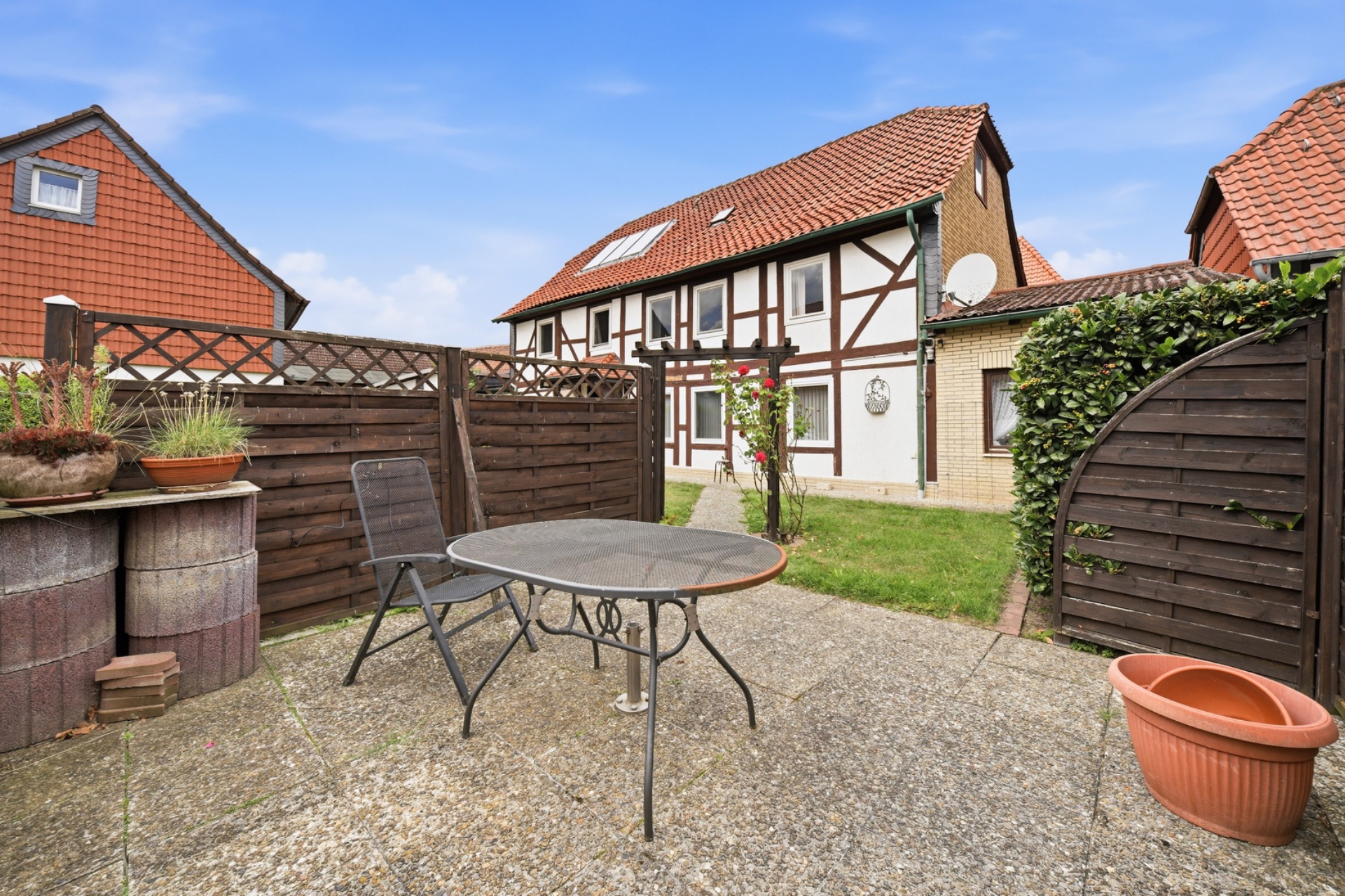 Gepflasterte Terrasse mit Tisch, Stuhl und Pflanzkübeln vor einem Fachwerkhaus mit rotem Dach.