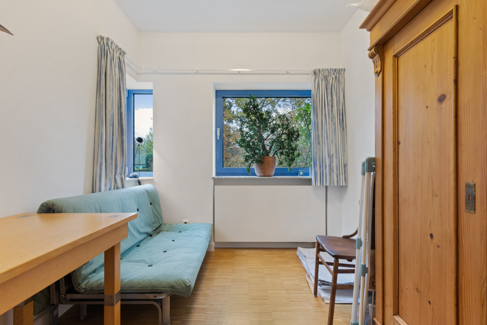 Kleines Zimmer mit grüner Schlafcouch, Holztisch, Stuhl, Kleiderschrank und Fenster mit Pflanze.