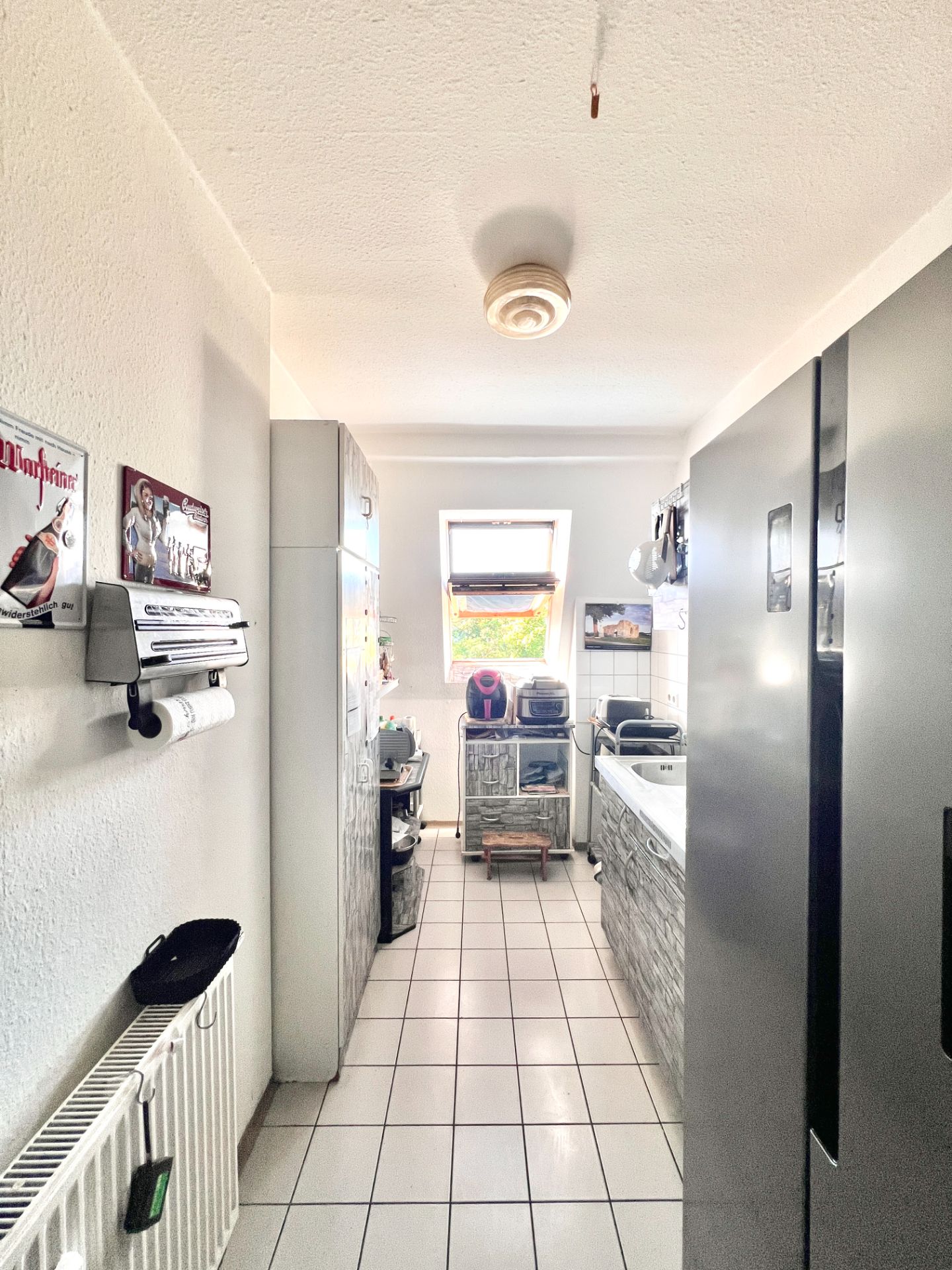 Schmale Küche mit Fliesenboden, dunklen Schränken und einem Fenster am Ende.