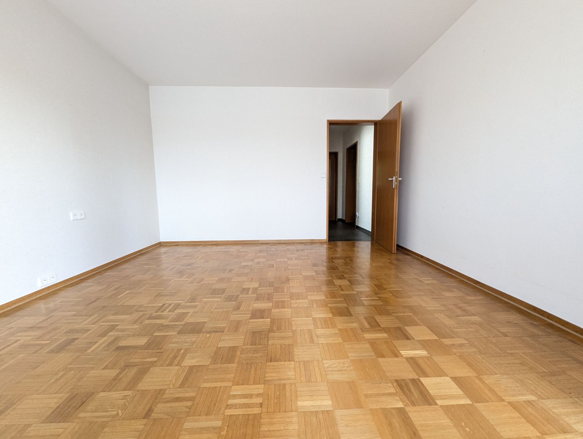 Leerer Raum mit Parkettboden, weißen Wänden und geöffneter Holztür.