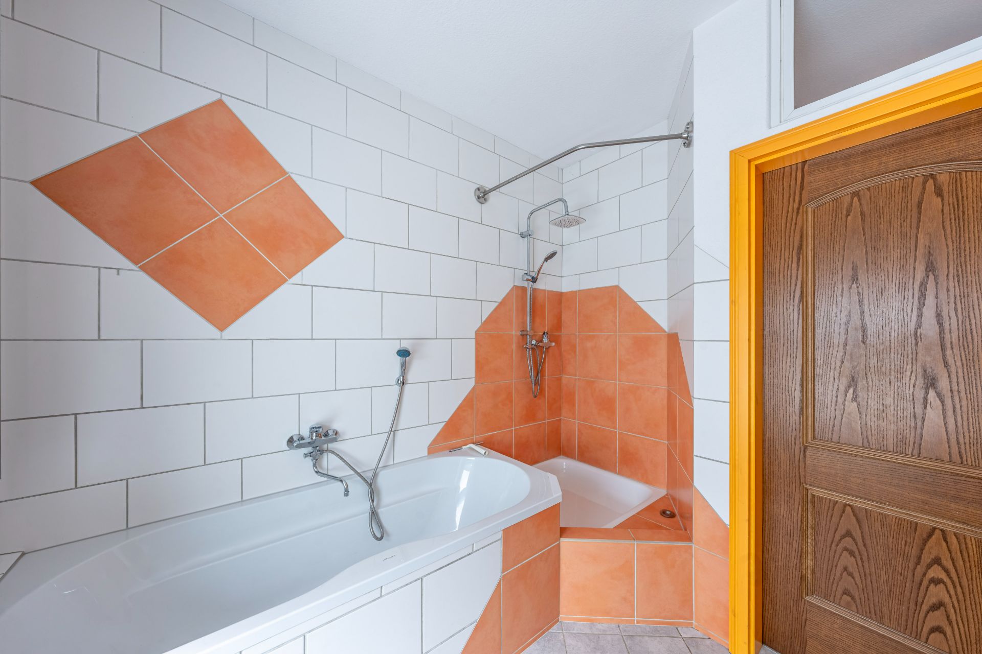 Badezimmer mit Badewanne, Dusche, weißen und orangefarbenen Fliesen und Holztür mit gelbem Rahmen.