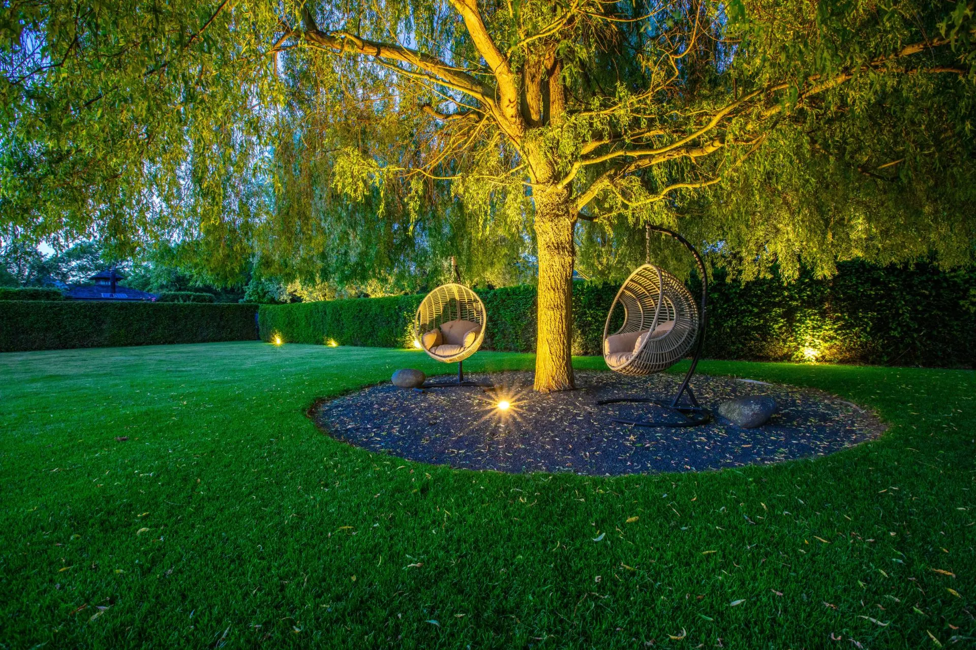 Im Garten stehen zwei runde Hängesessel unter einem großen Baum, beleuchtet von Bodenlichtern.