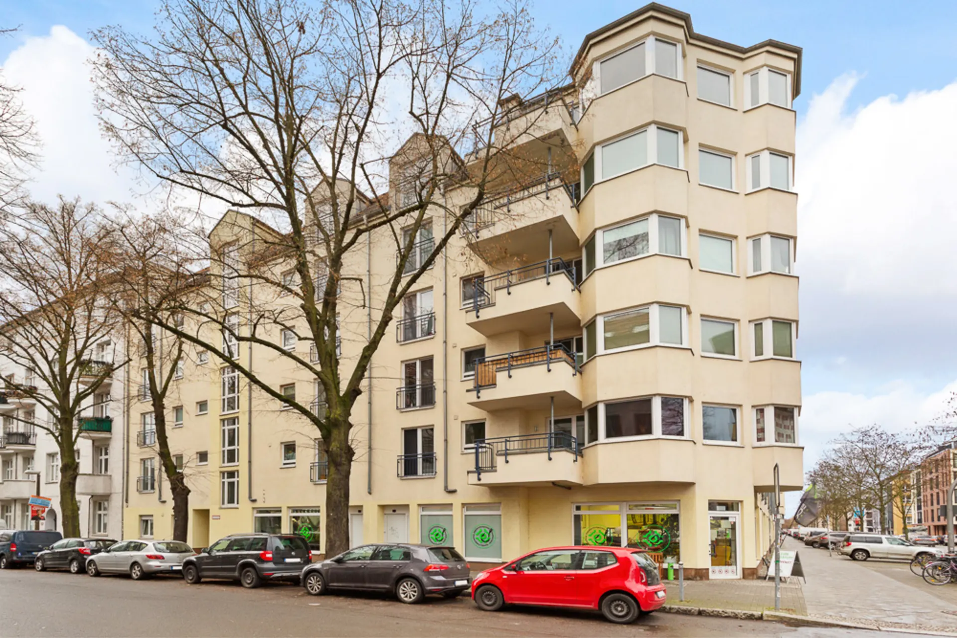Mehrstöckiges Wohngebäude mit Balkonen und großen Fenstern an einer Straßenecke, Autos parken davor.