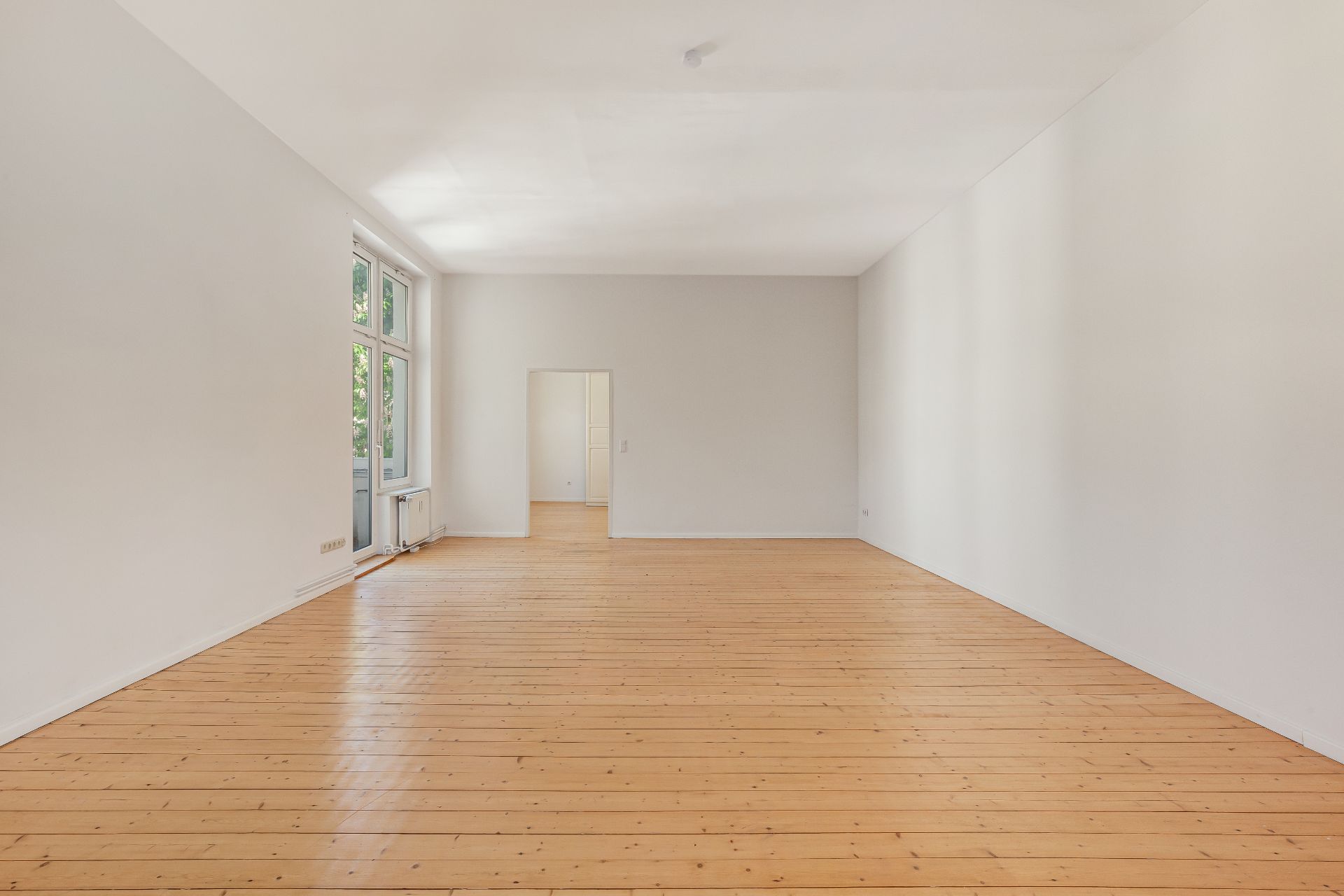 Heller, leerer Raum mit Holzboden, großen Fenstern und weiß gestrichenen Wänden.