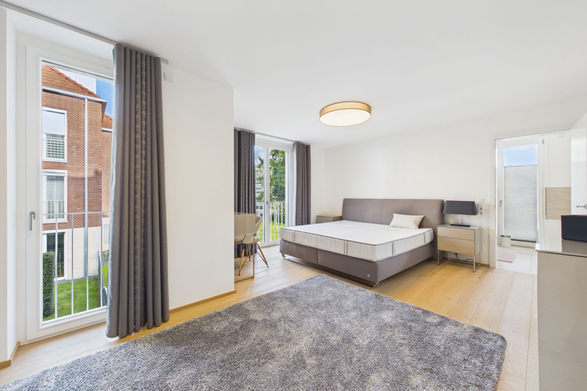 Helles Schlafzimmer mit großem Bett, Teppich, Schreibtisch und zwei bodentiefen Fenstern.