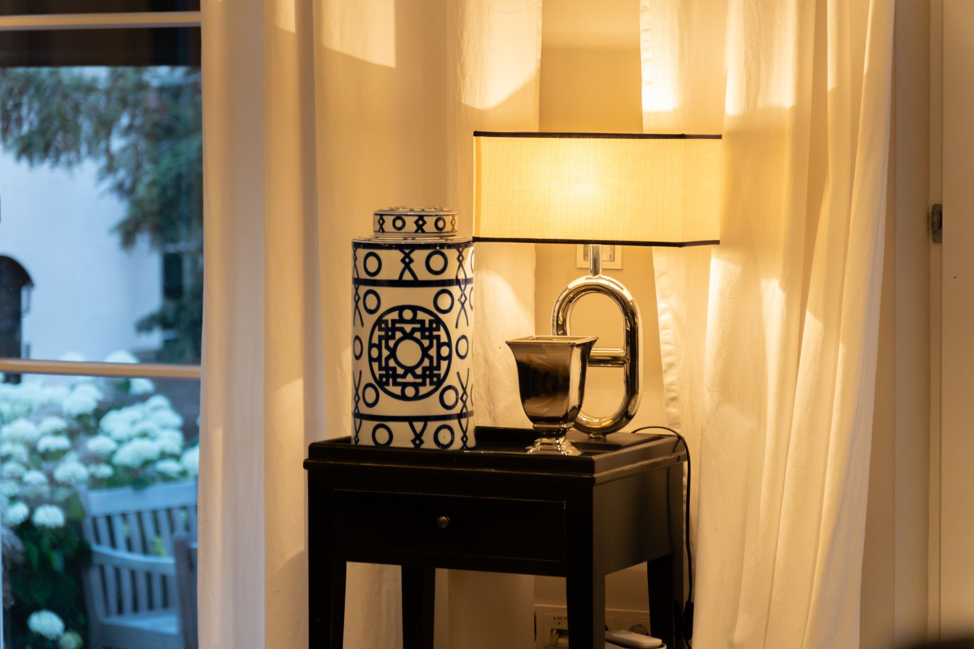 Tisch mit Lampe, Vase und Dekoration vor weißen Vorhängen in einem Wohnraum.