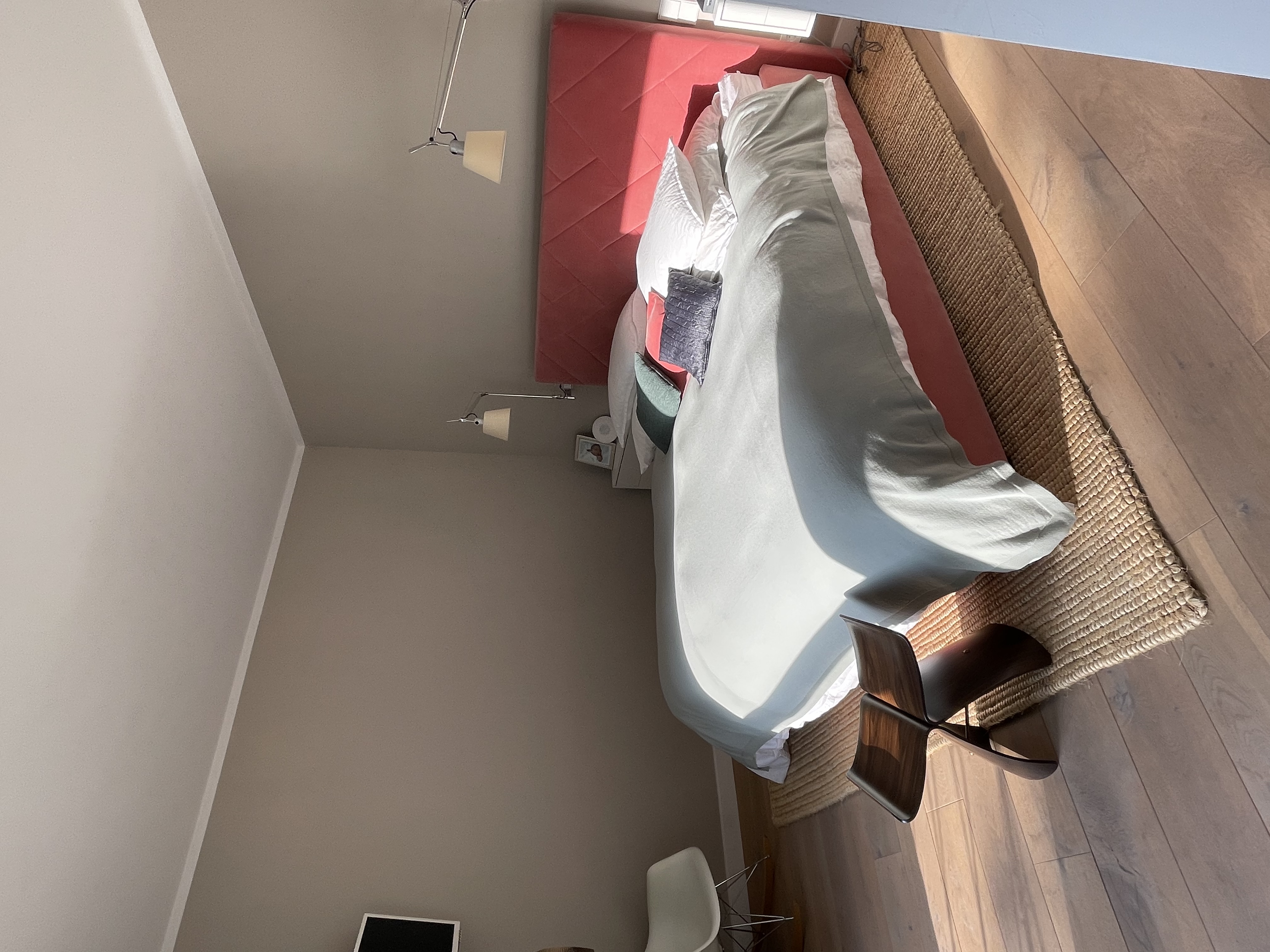 Helles Schlafzimmer mit Doppelbett, rosa Kopfteil, Holzstuhl und Teppich auf Holzboden.