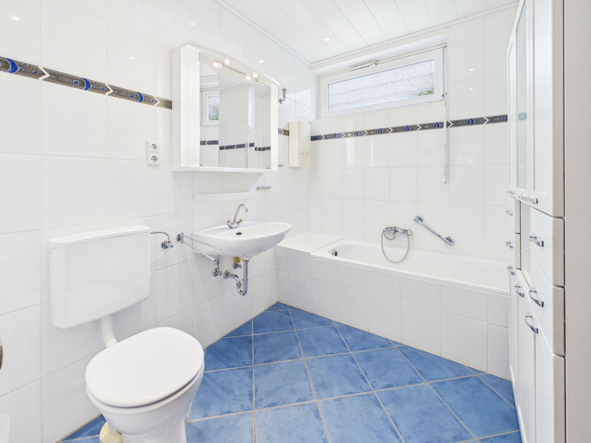 Helles Badezimmer mit Badewanne, Waschbecken, Toilette und blau gefliestem Boden.