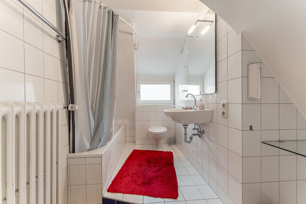 Helles Badezimmer mit Badewanne, Waschbecken, WC und rotem Teppich auf weißen Fliesen.
