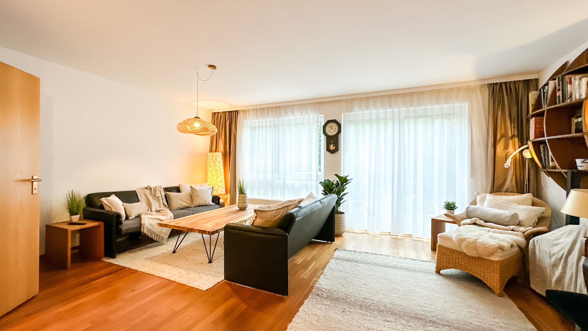 Helles Wohnzimmer mit Sofas, Couchtisch, Sessel, Regal und großen Fenstern mit Vorhängen.