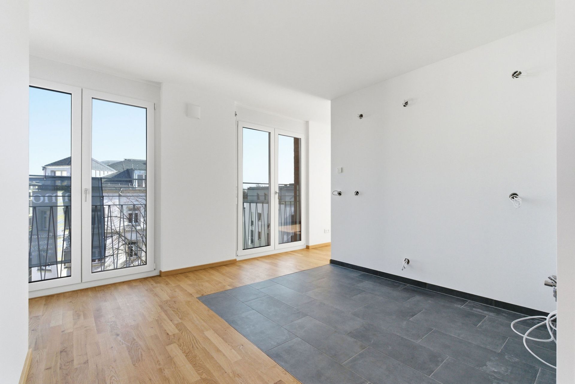 Heller leerer Raum mit Parkettboden, Fliesenbereich und zwei bodentiefen Fenstertüren.