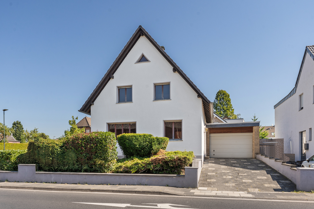 Weißes Einfamilienhaus mit hoher Giebeldach und Garage, umgeben von Hecken und Vorgarten.