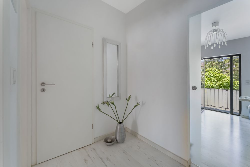 Heller Flur mit weißer Tür, Spiegel und Vase mit Blumen, Blick in ein Zimmer mit Balkon.