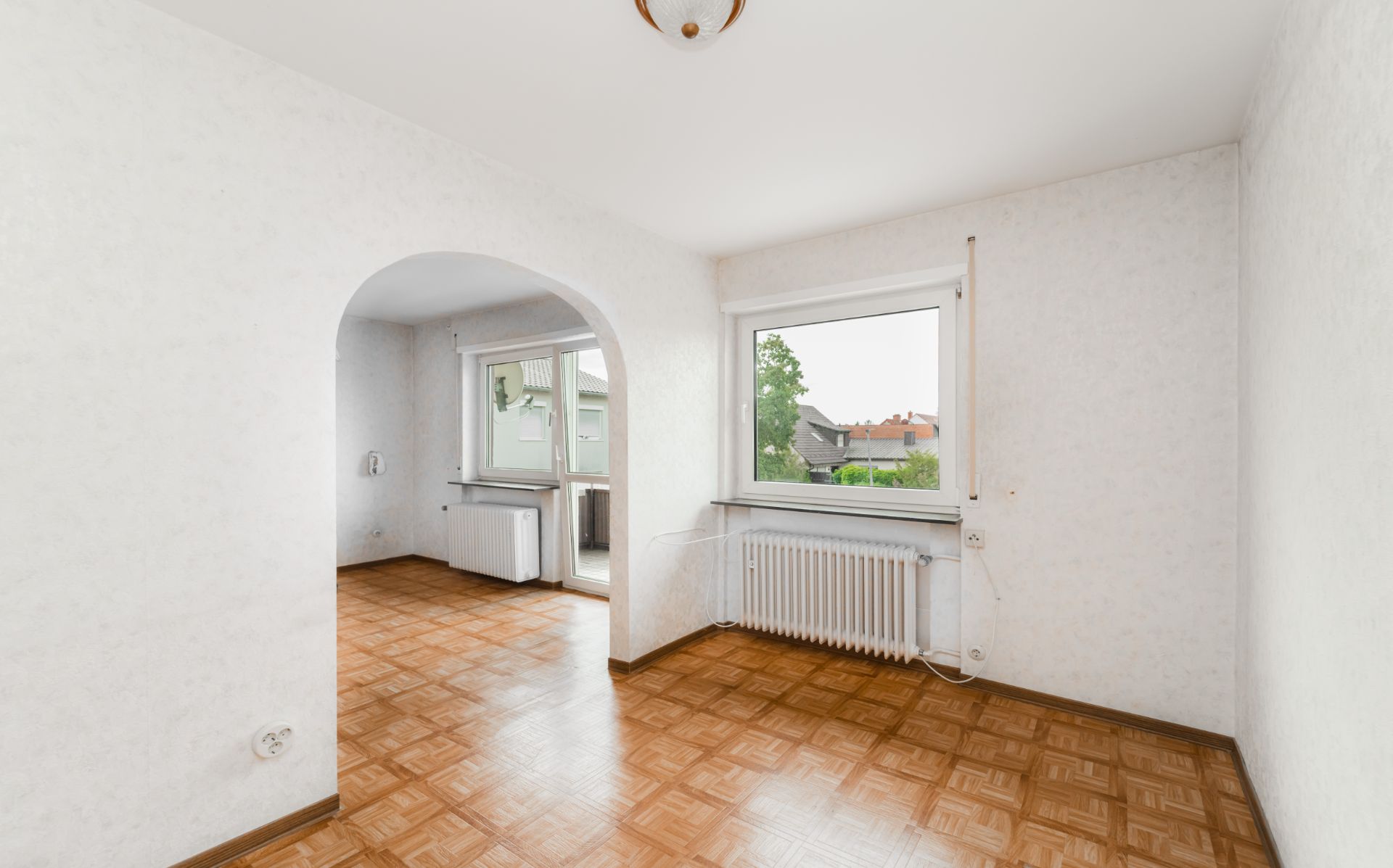 Heller Raum mit Parkettboden, großem Fenster und Rundbogen zu einem weiteren Zimmer.