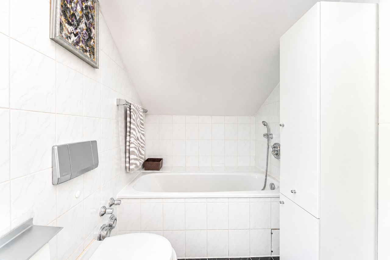 Helles Badezimmer mit Badewanne, Toilette und weißem Schrank unter einer Dachschräge.