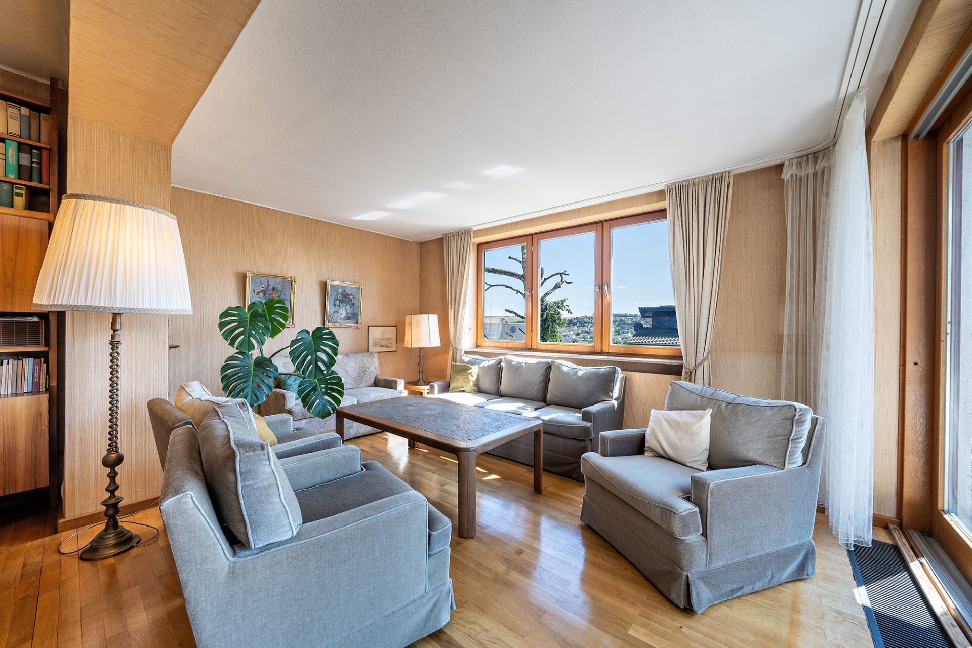 Helles Wohnzimmer mit grauen Sofas, Sessel, großem Tisch, Holzboden und großen Fenstern.